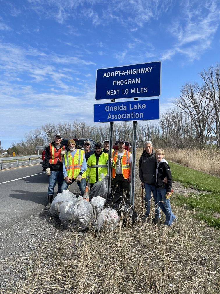 Blue Adopt-a-highway program Oneida Lake Association signs with white letters. Oneida Lake Association members standing underneath with bags full of garbage April 2026